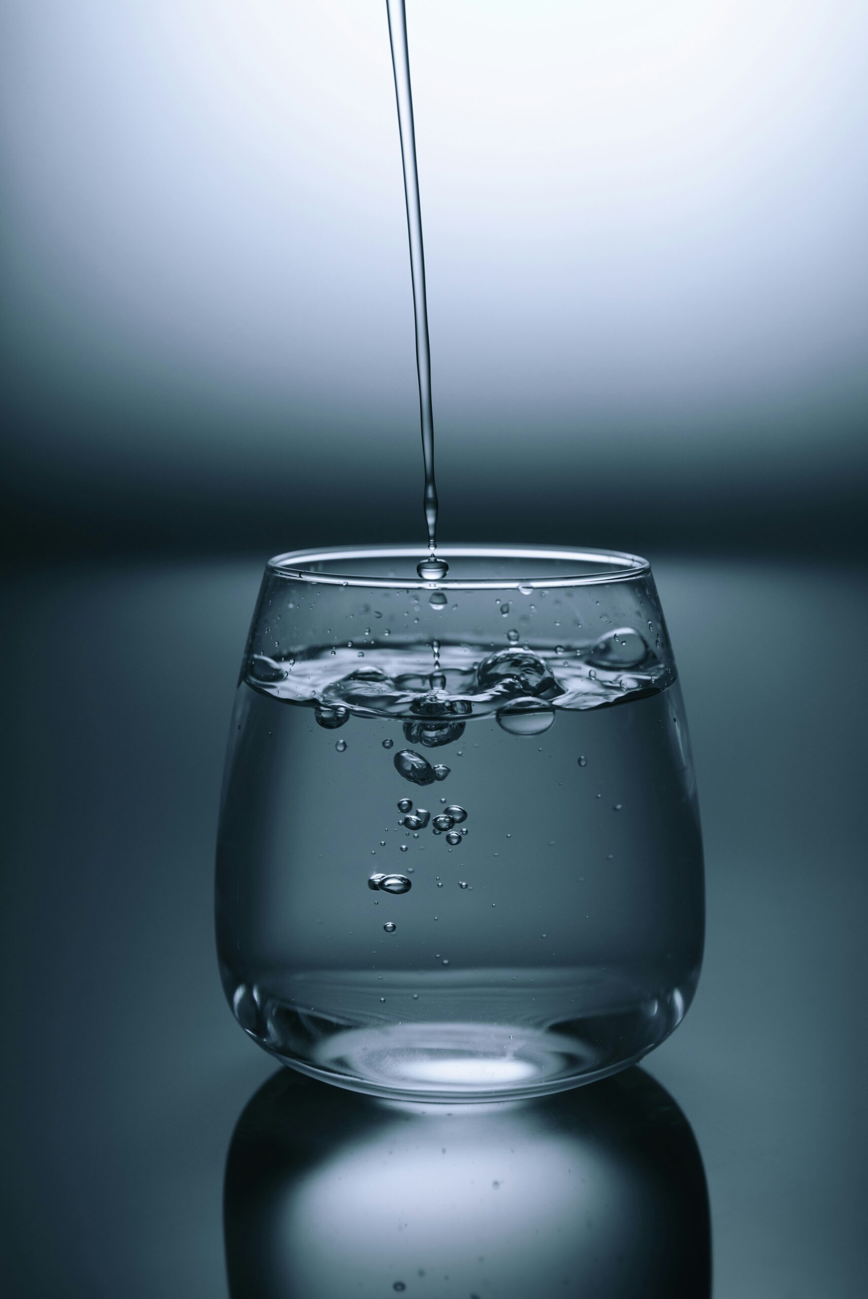 A studio shot of water being poured in a steady stream into a clear, curved glass. The water is splashing and creating bubbles inside the glass against a moody, dark grey background.