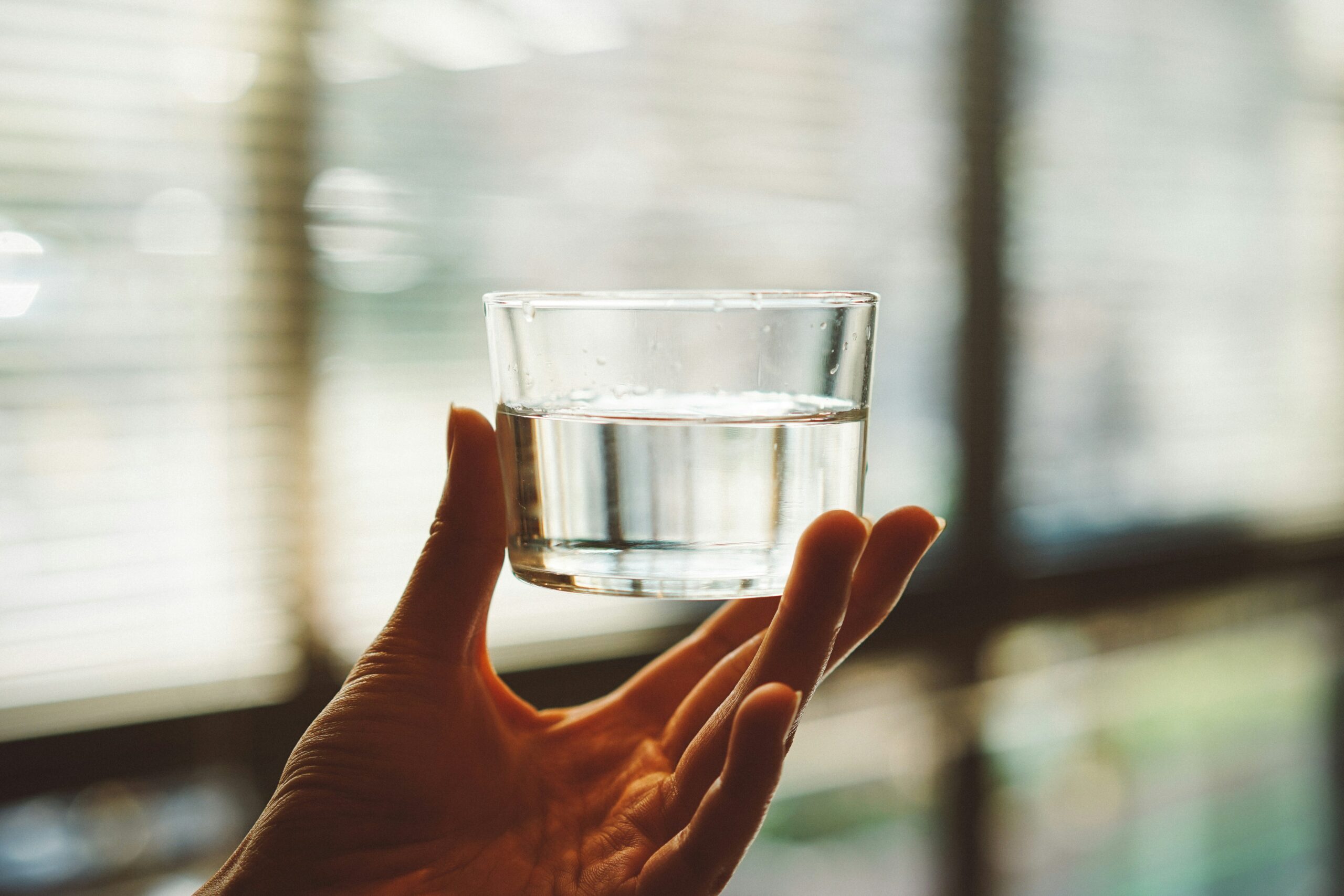 A person’s hand holding a small, clear glass half-filled with water. The image is shot from a low angle with soft, natural light filtering through window blinds in the background.
